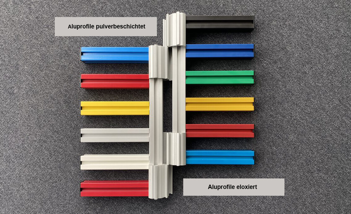 Bunte Aluminiumprofile in verschiedenen Farben liegen auf einem grauen Hintergrund, einige sind pulverbeschichtet, andere eloxiert.