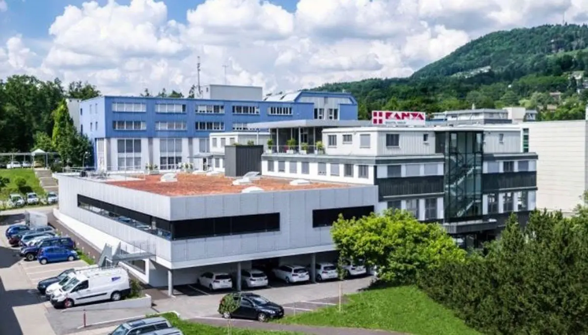 Das moderne KANYA-Gebäude in der Schweiz mit blauer Fassade und großen Fenstern, umgeben von Bäumen und einem Parkplatz voller Autos.